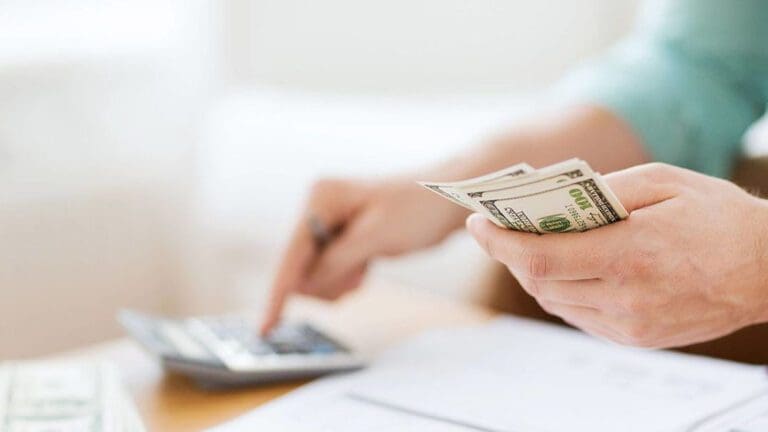 Hands shown over desk with paperwork. One hand is working a calculator and the other is holding cash.