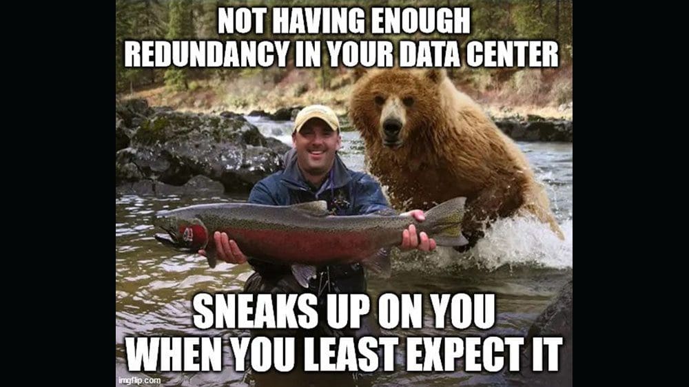 Man in river holding large fish poses for picture. He doesn't realize a bear is behind him ready to take the fish. Text reads Not having enough redundancy in your data center sneaks up on you when you least expect it.